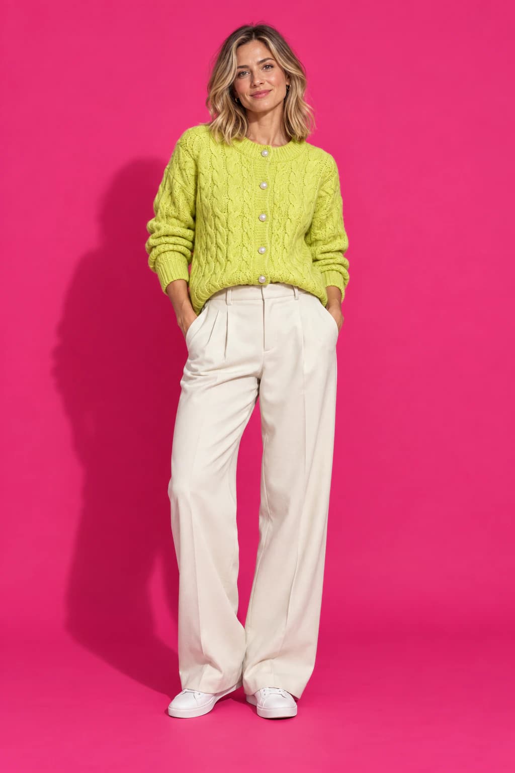 Model in a chunky chartreuse cable-knit cardigan and cream trousers on a fuchsia backdrop