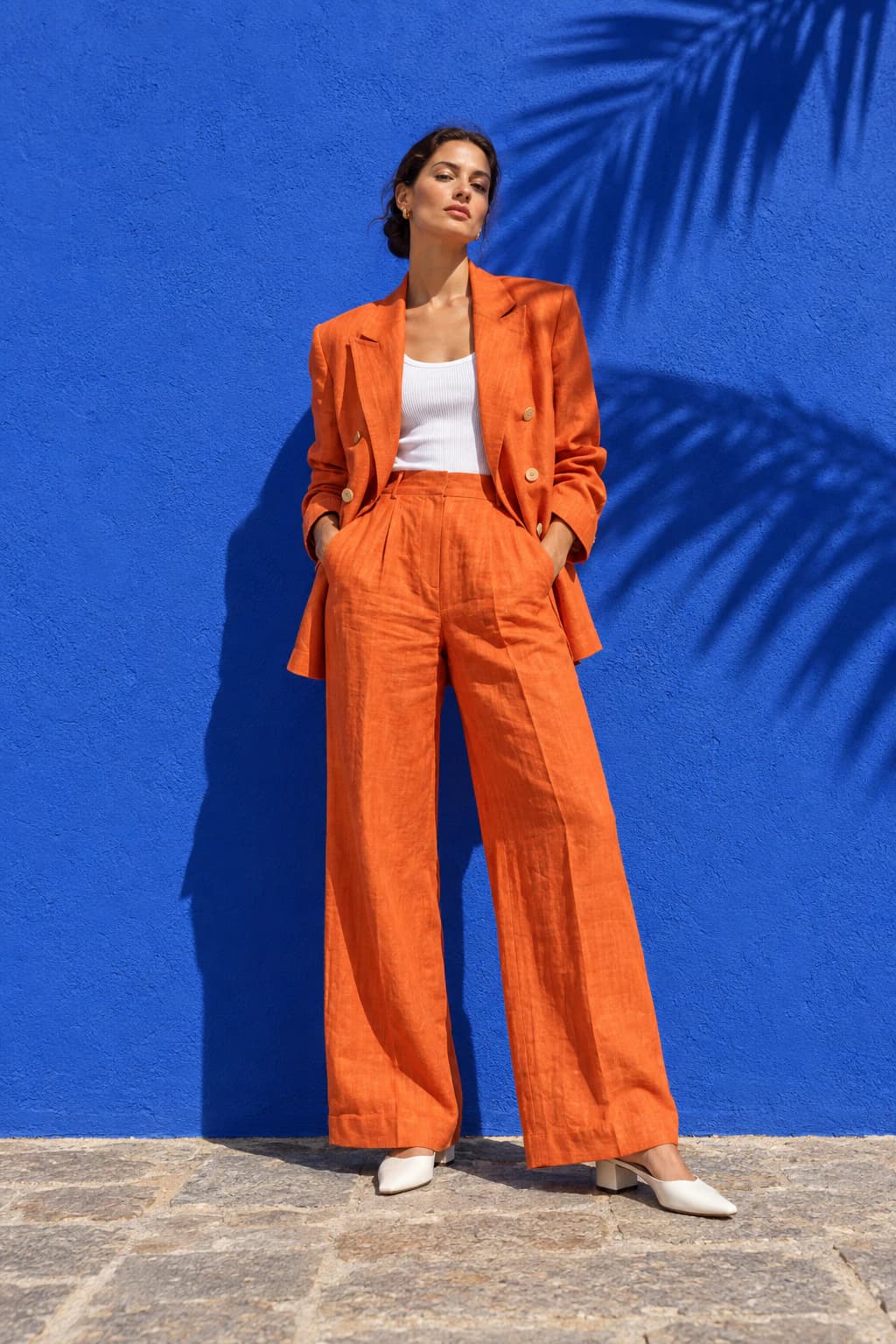 Model in an oversized tangerine linen suit against a cobalt wall with palm shadows