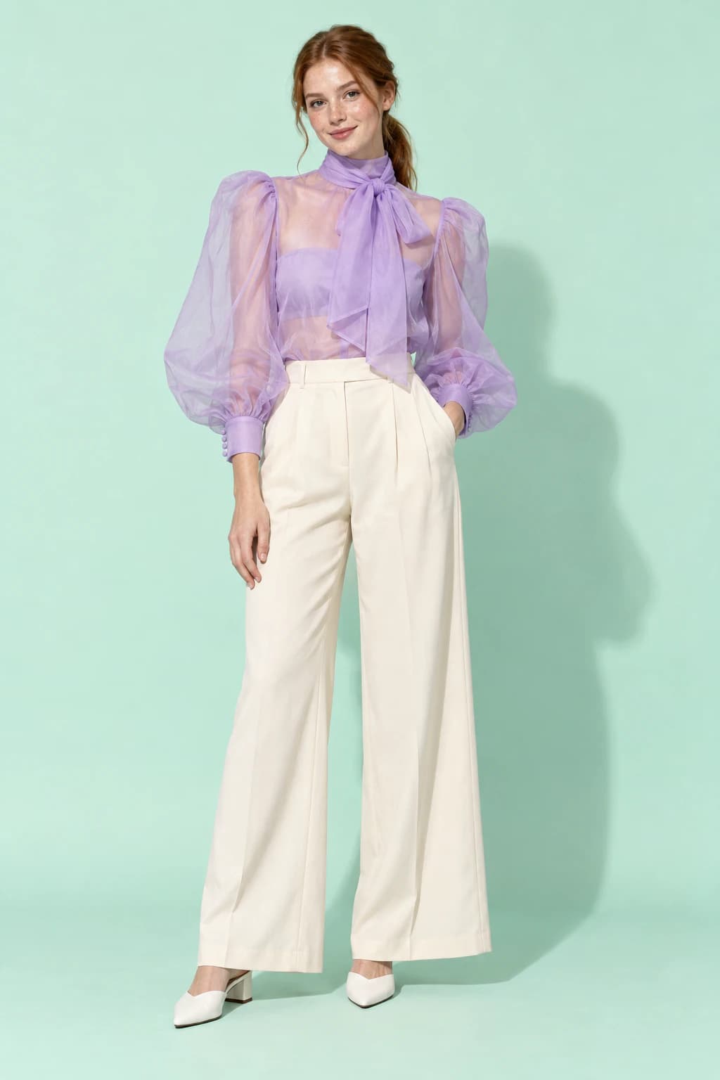 Model in a sheer lilac organza puff-sleeve blouse and cream trousers on a mint backdrop